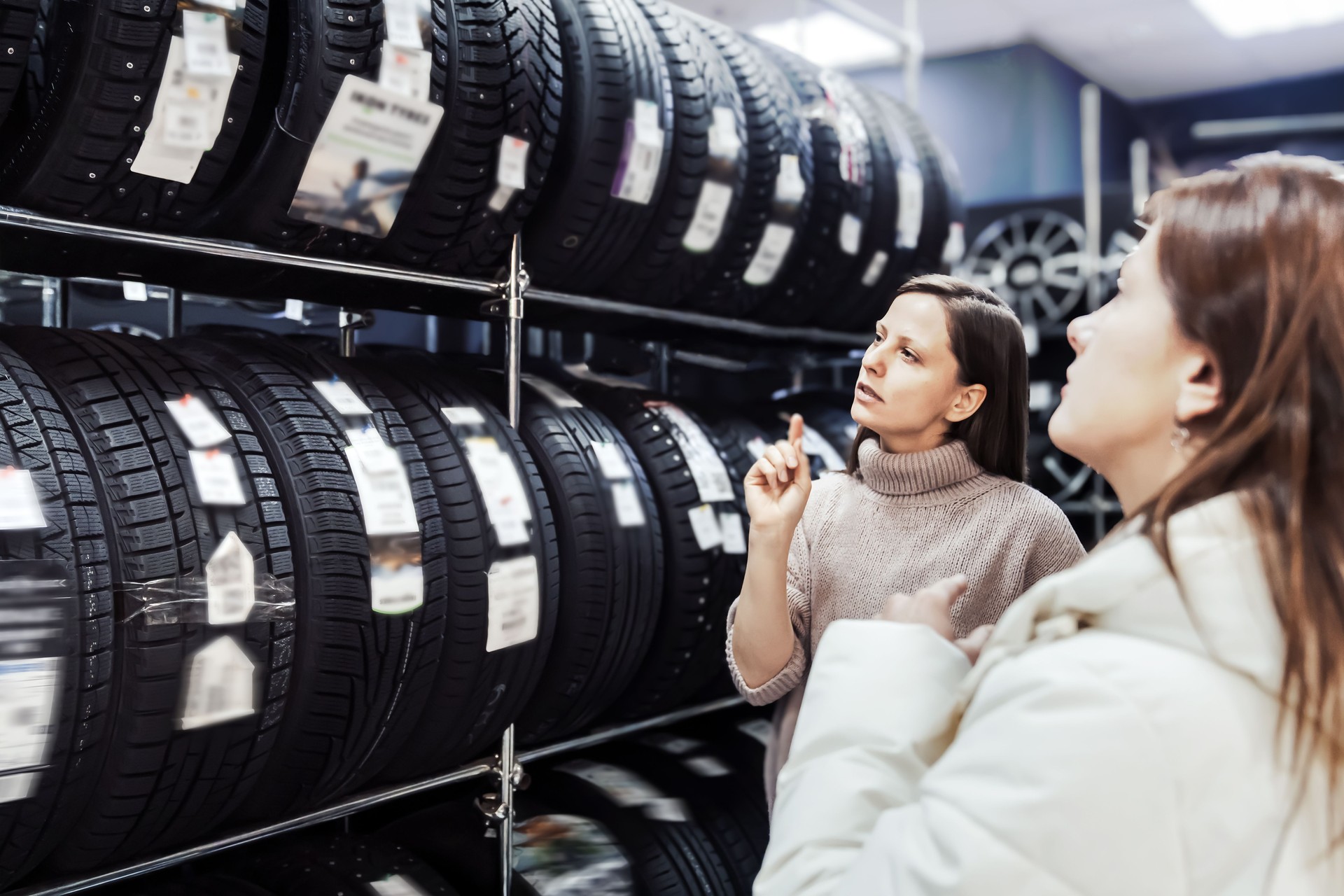 Two women engage in consultation at tire shop, discussing best options for seasonal tire purchases. One woman thoughtfully examines available tires displayed on shelves while seller provides guidance